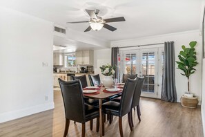 A dining area with a table set for four, a ceiling fan, and a view of the kitchen and outside.