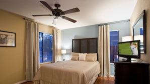 Un dormitorio con cama, ventilador de techo, televisión y ventana con cortinas.