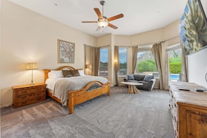 A bedroom with a bed, a nightstand, a dresser, a ceiling fan, a window with curtains, and a view of greenery outside. - Resort Style Pool Home- Troon - Updated (Scottsdale)