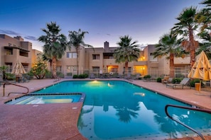 A swimming pool with lounge chairs and umbrellas, surrounded by palm trees and a building with balconies.