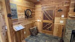 Interior de una cabaña de madera con suelo de baldosas de piedra, una puerta de madera, un espejo y una pequeña mesa con un letrero.