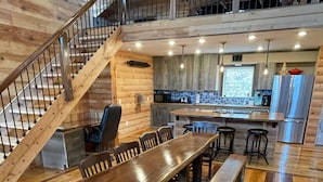 Un comedor amplio con una larga mesa de madera y sillas, una escalera que sube a un nivel superior y una cocina con electrodomésticos modernos.