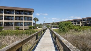 A wooden boardwalk leading to a multi-story building with balconies, surrounded by vegetation and clear skies. - Peek of the ocean King size bed just Steps from the Beach (Hilton Head Island)