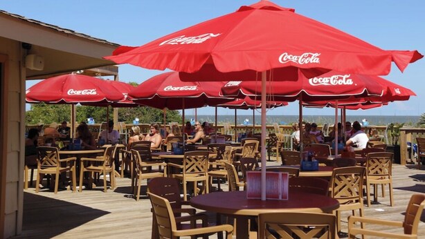 An outdoor seating area with wooden tables and chairs, red Coca-Cola branded umbrellas, and a clear view of the ocean. - Peek of the ocean King size bed just Steps from the Beach (Hilton Head Island)