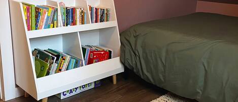 A room with a white bookshelf filled with books, a green bed, and a woven rug on the floor.
