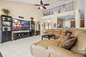 A spacious living room with a large flat-screen TV, a leather sofa, and a dining area with a kitchen in the background.