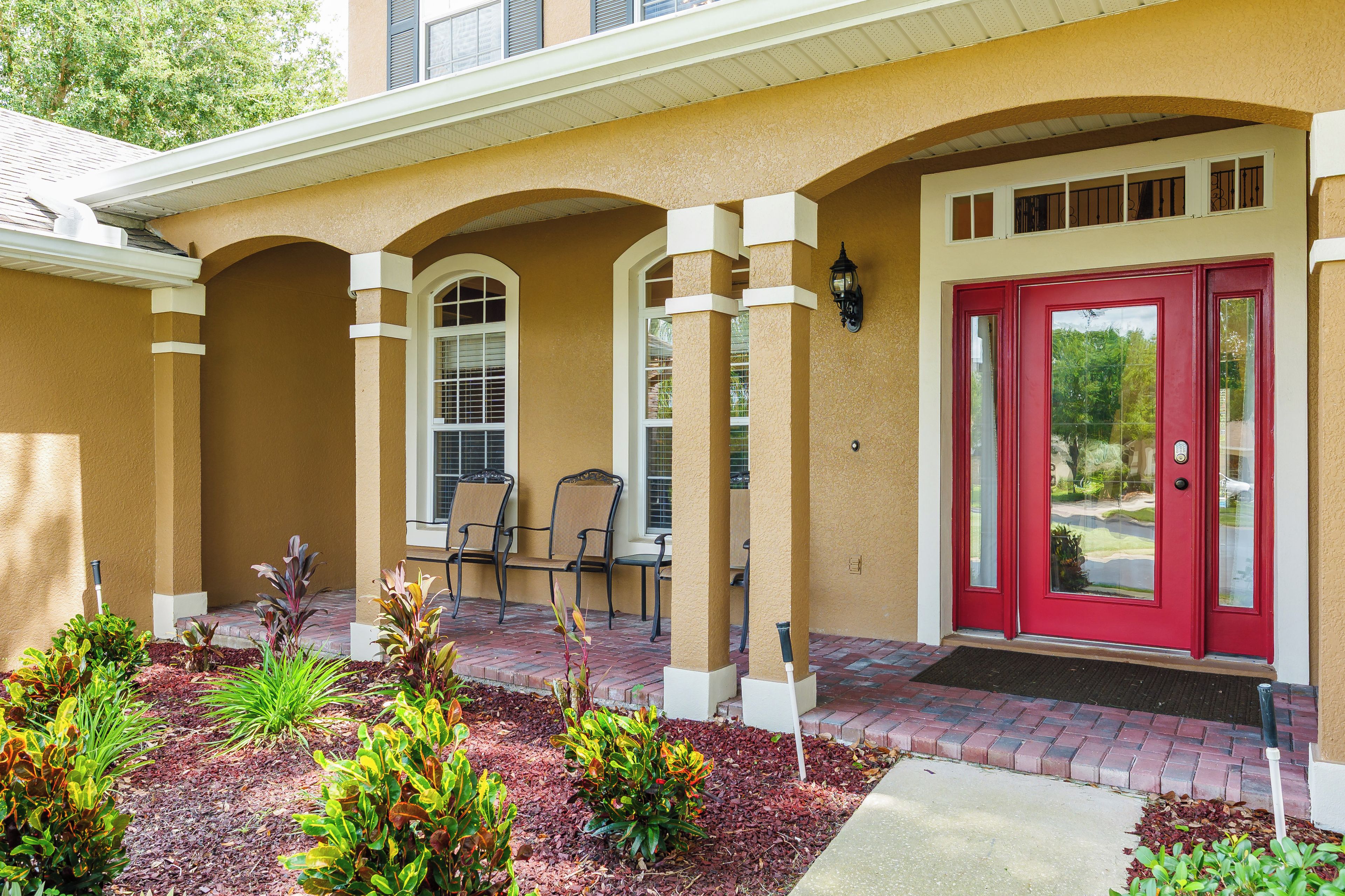 A building entrance with a red door, a covered patio area with chairs, and a landscaped garden.