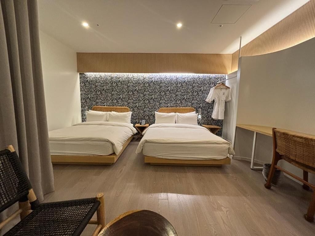 A hotel room with two beds, a wooden floor, a wall with a leaf pattern, a bench, and a hanging garment rack.