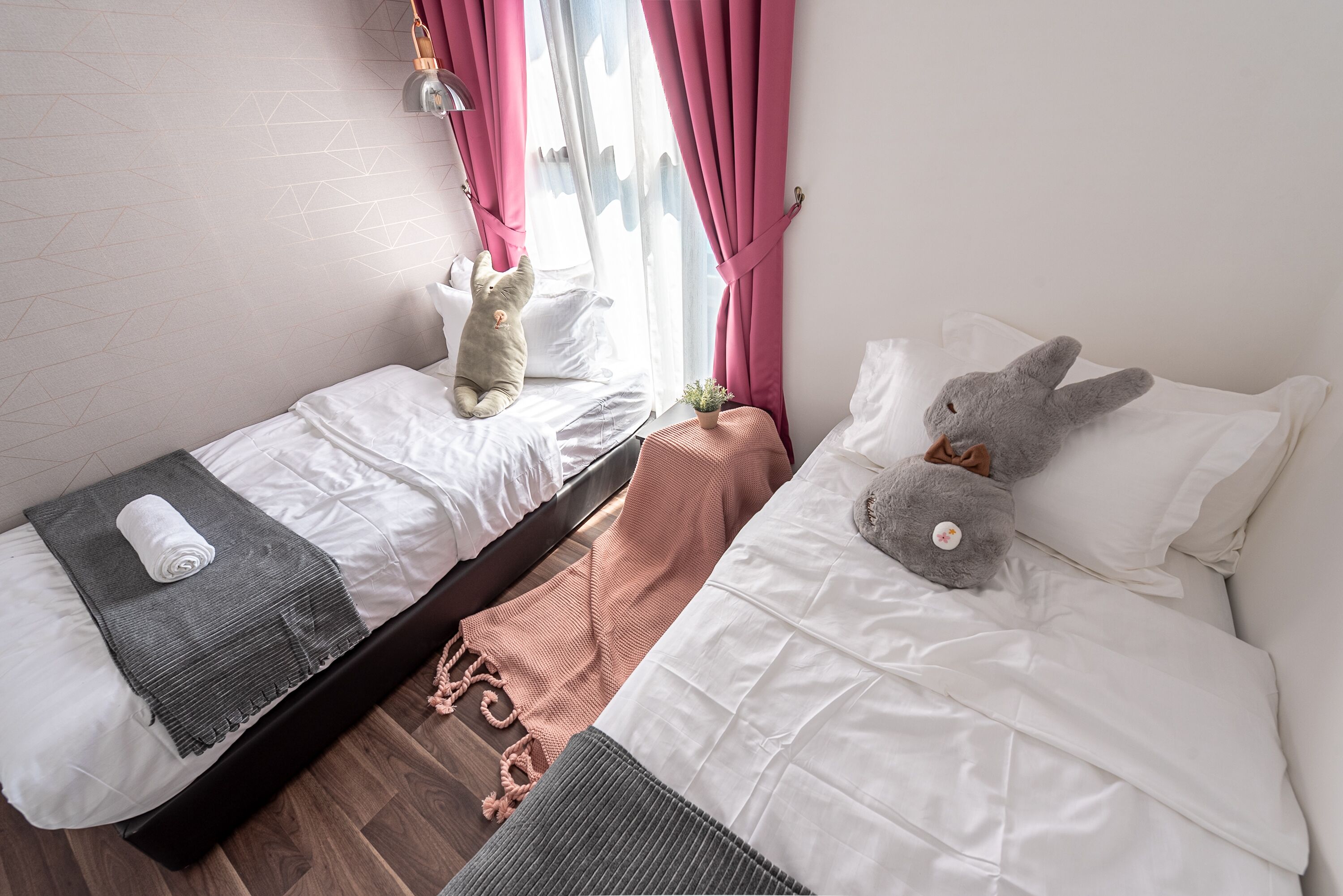 A bedroom with two beds, a window with pink curtains, a small potted plant, and a teddy bear on one bed.