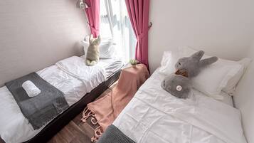A bedroom with two beds, a window with pink curtains, a small potted plant, and a teddy bear on one bed.