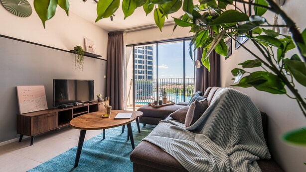 A modern living room with a sofa, coffee table, TV, and a balcony with a view of buildings. - 3BR Homestay with Pool View Bukit Rimau (Shah Alam)