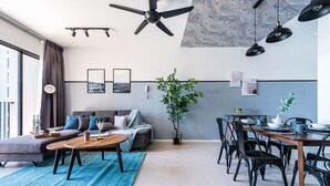 A modern living room with a sofa, coffee table, dining table, chairs, ceiling fans, pendant lights, and a large window. - 3BR Homestay with Pool View Bukit Rimau (Shah Alam)