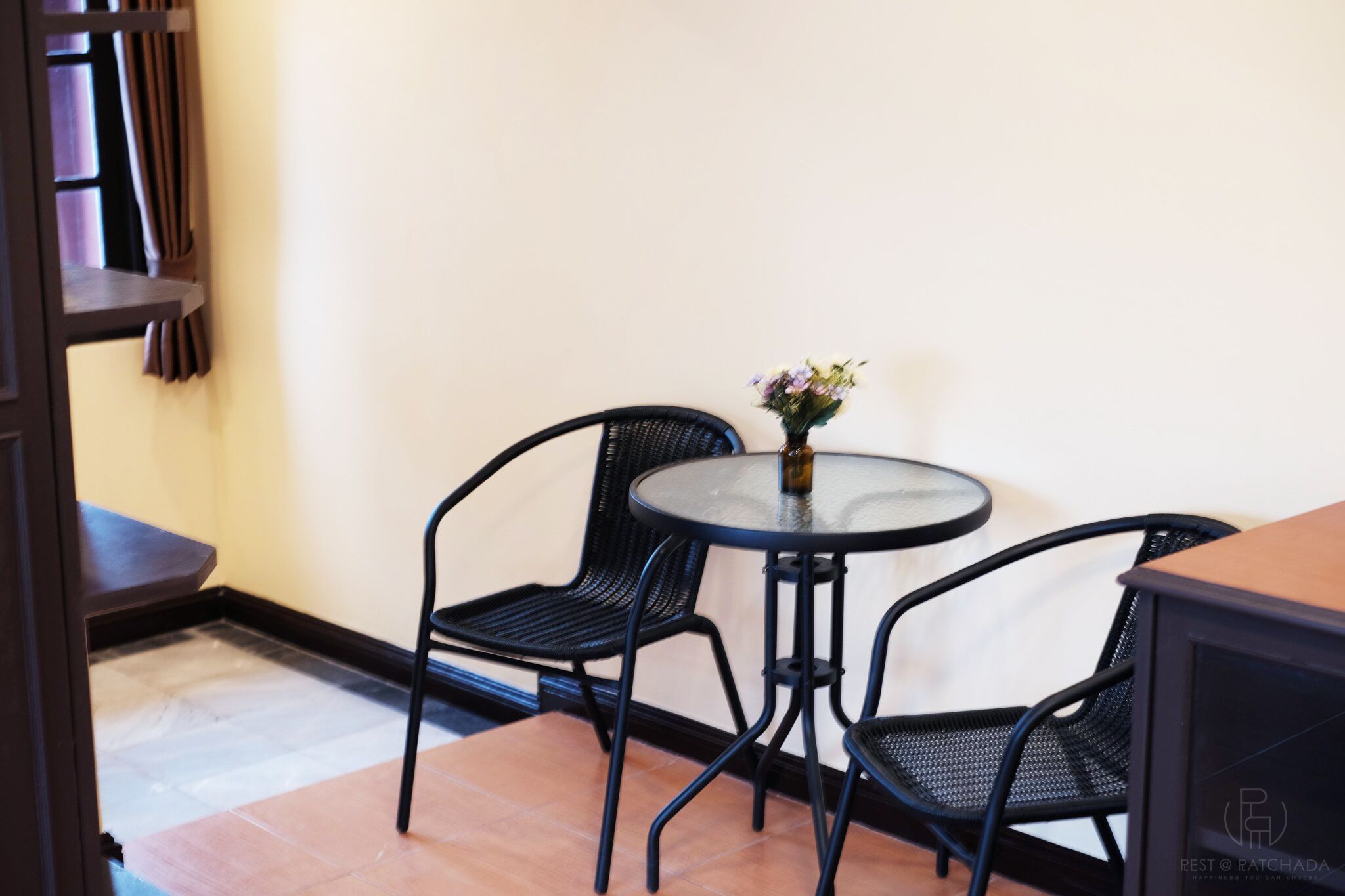 A small, enclosed room with a round glass table and two black chairs. A small vase with flowers is on the table.