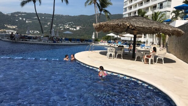 Una piscina con sillas de descanso y sombrillas, rodeada de palmeras y un edificio al fondo.