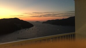 Vista al atardecer desde un balcón con vistas a un cuerpo de agua y colinas lejanas.