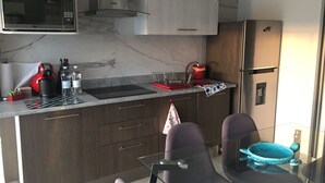 Cocina compacta equipada con microondas, hervidor de agua y dispensador de agua. Cuenta con un pequeño comedor con una mesa de vidrio y dos sillas.