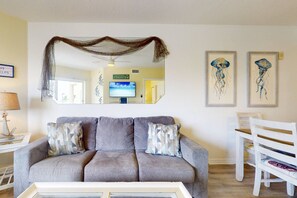 A living room with a grey sofa, two patterned throw pillows, a white coffee table, a white chair, and two framed artworks on the wall.