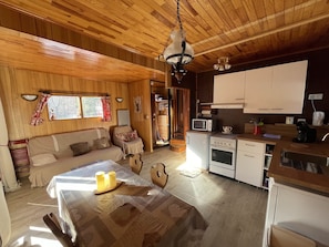 A cozy cabin interior with a wooden ceiling, a dining table with candles, a sofa, a kitchen area with a microwave and stove, and a window with curtains.