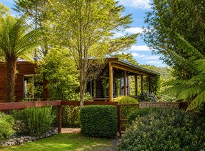 Una cabaña de madera con porche cubierto, rodeada de abundante vegetación y un letrero que dice 'CountryCottage'.
