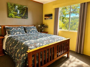 Ein Schlafzimmer mit einem hölzernen Bett, einem Fenster mit Vorhängen, einem Gemälde an der Wand und einem Nachttisch mit Lampe und Blumen.