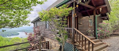 A rustic log cabin with a stone staircase leading to a wooden porch, surrounded by lush greenery and a scenic view of a lake.