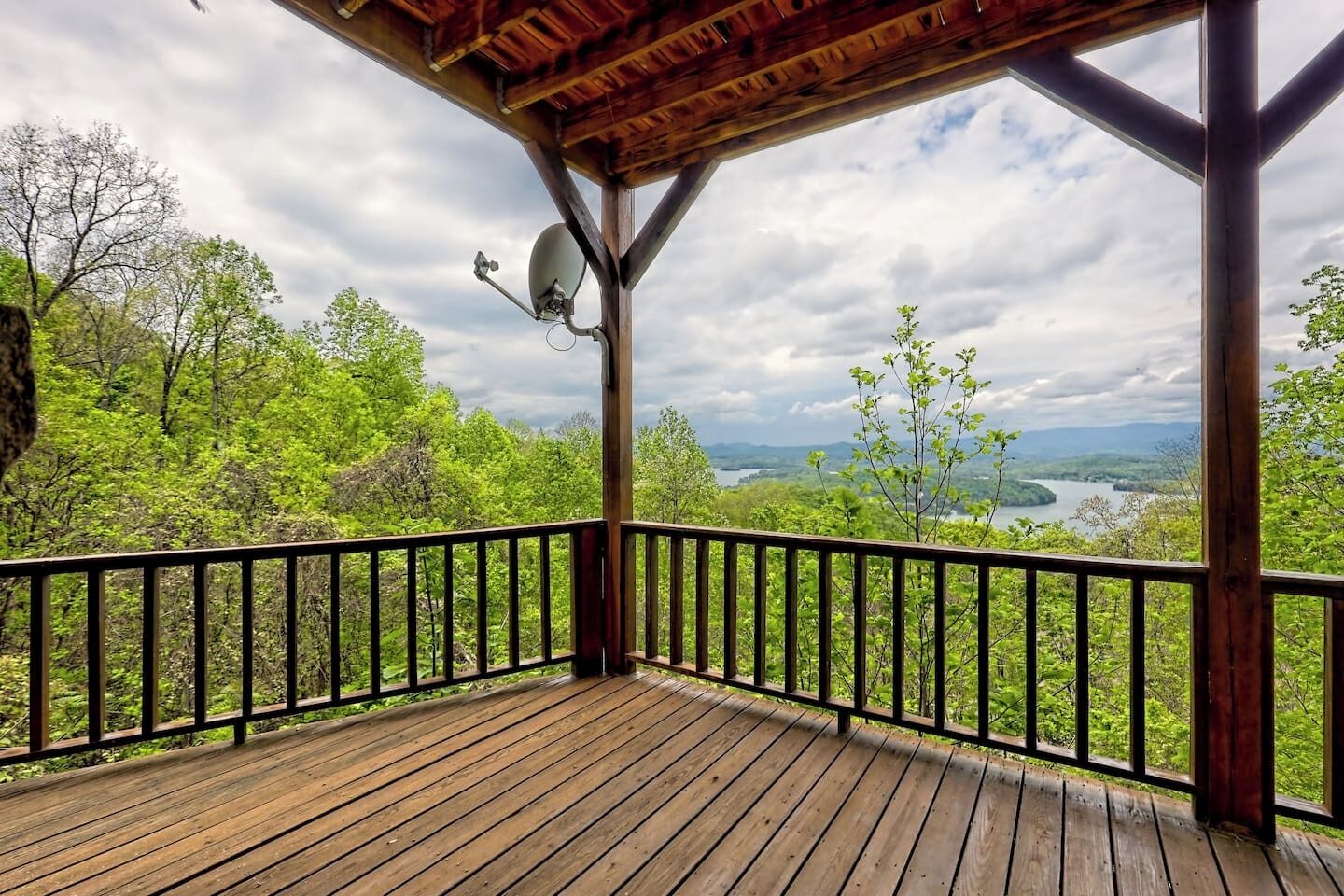 A wooden deck with a metal railing, a satellite dish, and a view of a lake surrounded by trees.