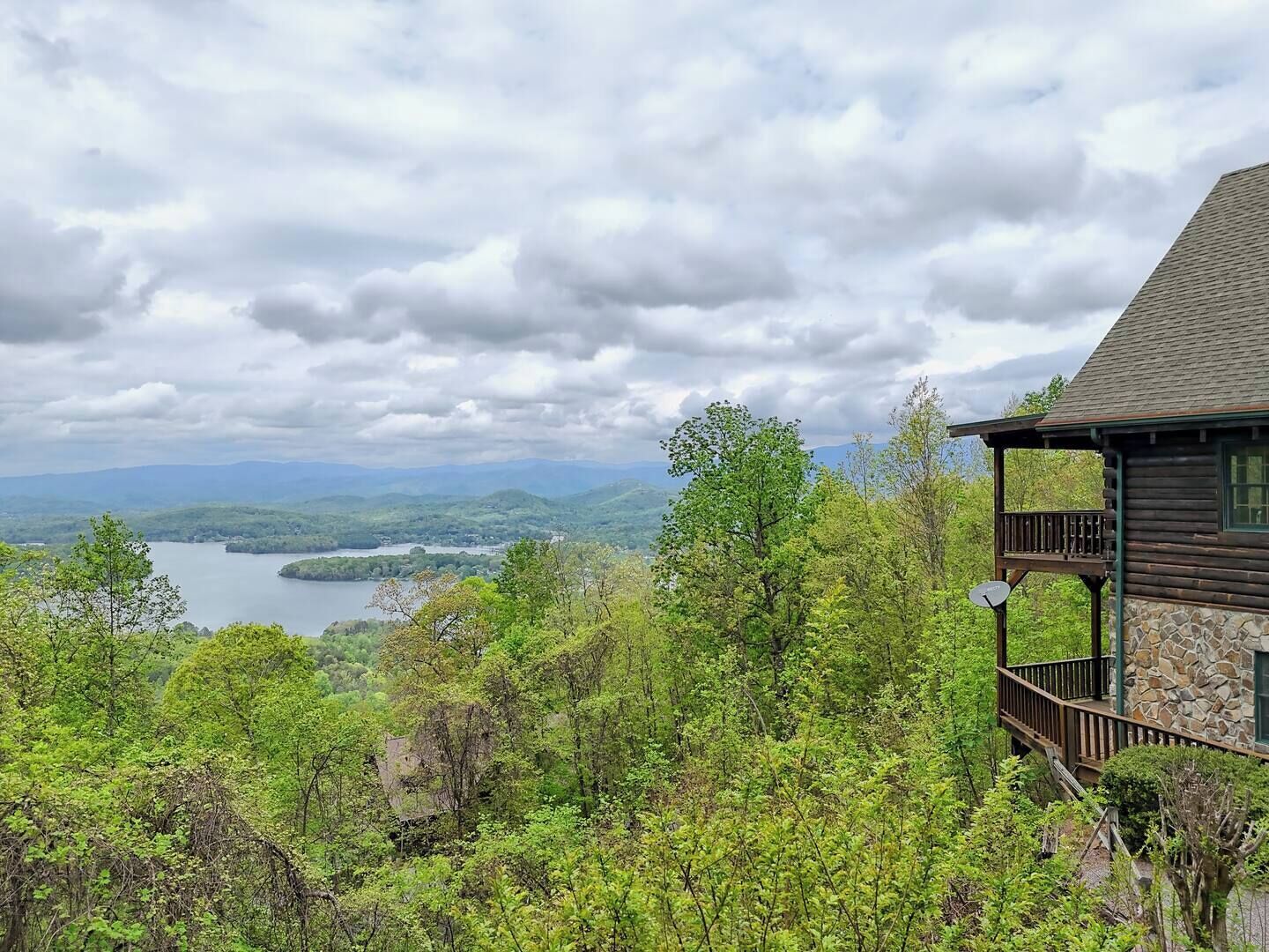 Una cabaña de fachada de piedra con balcón con vistas a un lago y montañas boscosas.