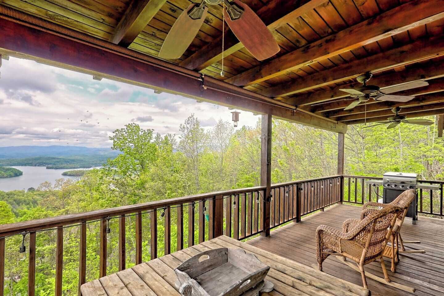 A wooden deck with a view of a lake and mountains, a grill, and wicker chairs.