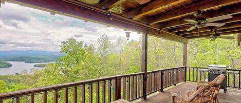 A wooden deck with a view of a lake and mountains, a grill, and wicker chairs.