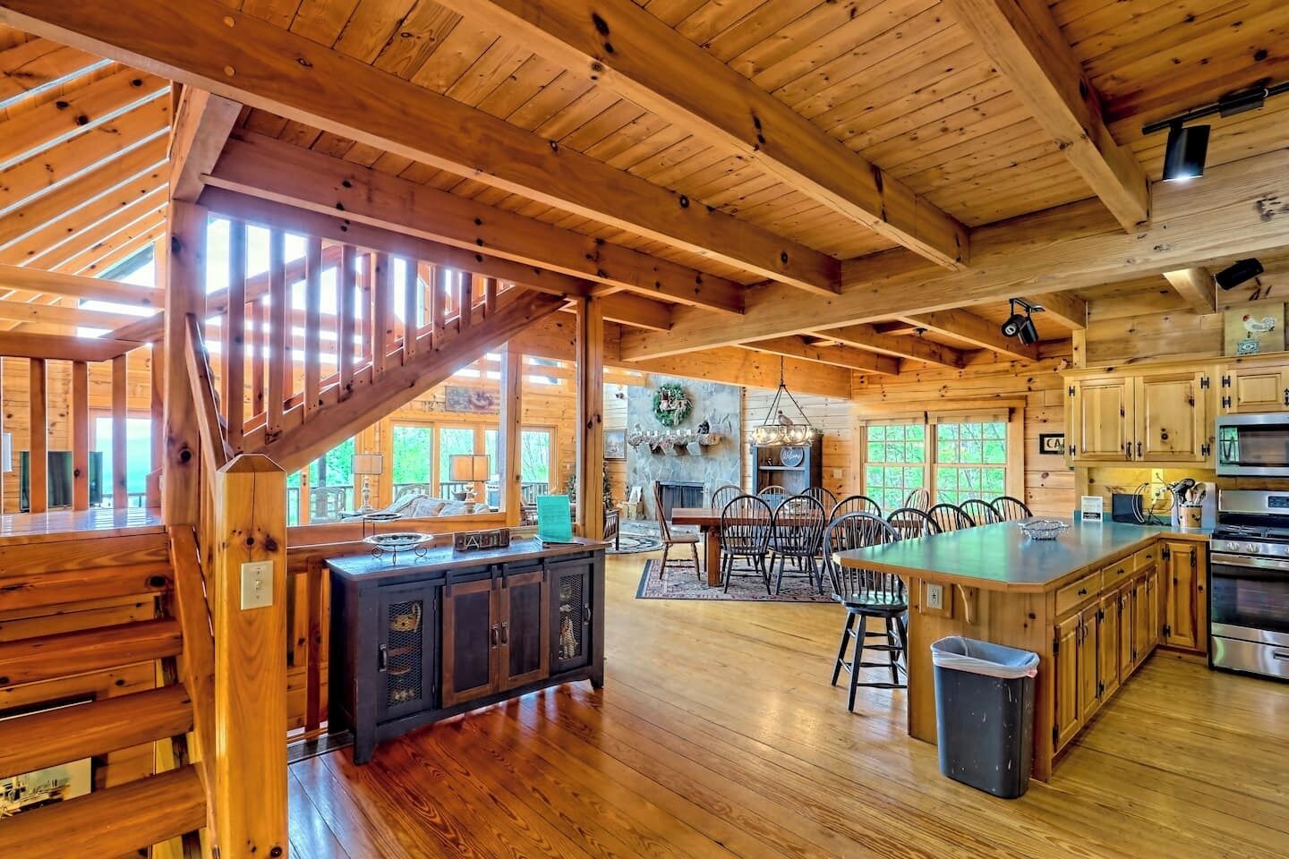 Un interior espacioso de cabaña de madera con un amplio comedor, cocina y una escalera que lleva a un nivel superior.
