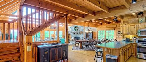 Un interior espacioso de cabaña de madera con un amplio comedor, cocina y una escalera que lleva a un nivel superior.