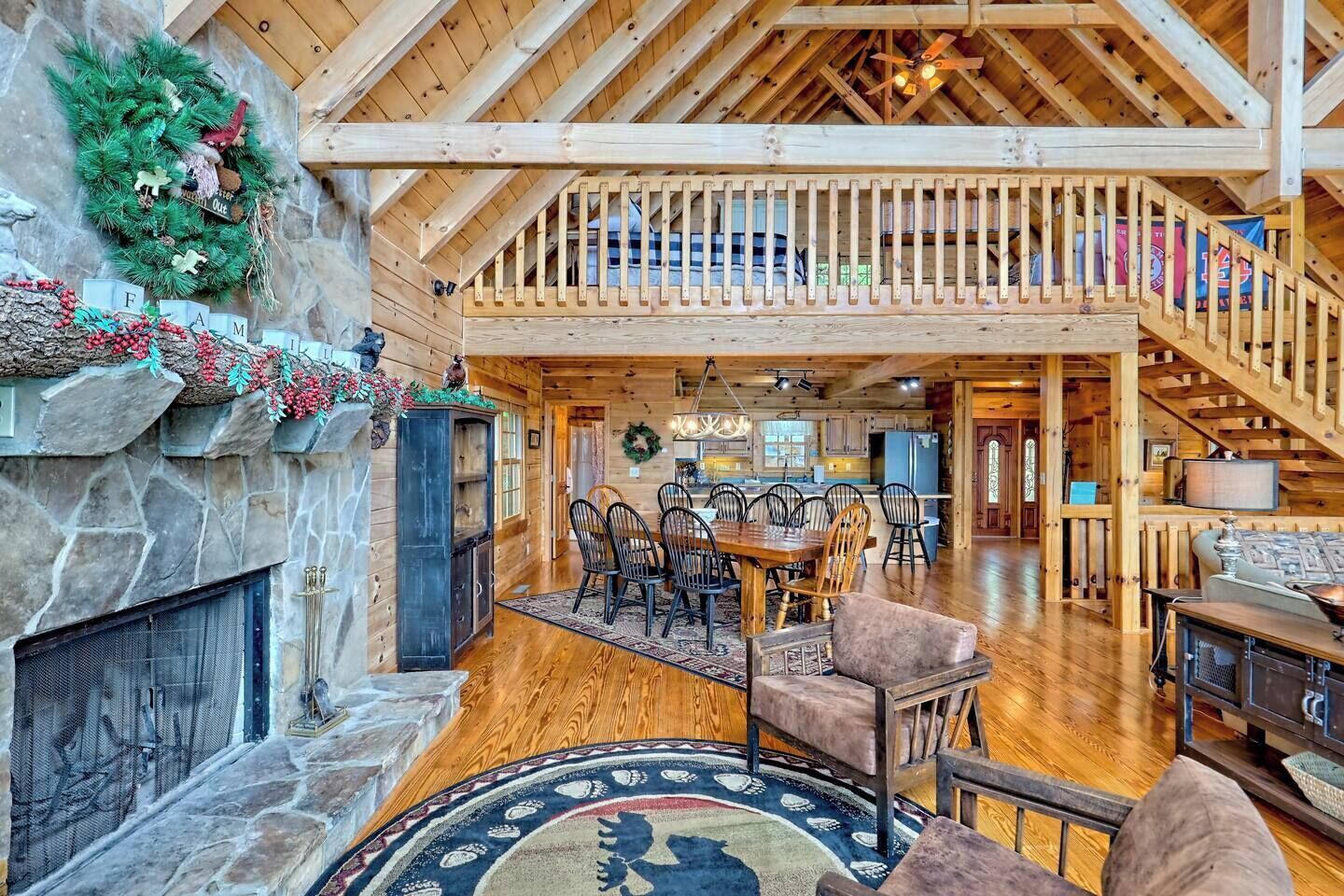 A spacious log cabin interior with a stone fireplace, a dining area, and a staircase leading to an upper level.