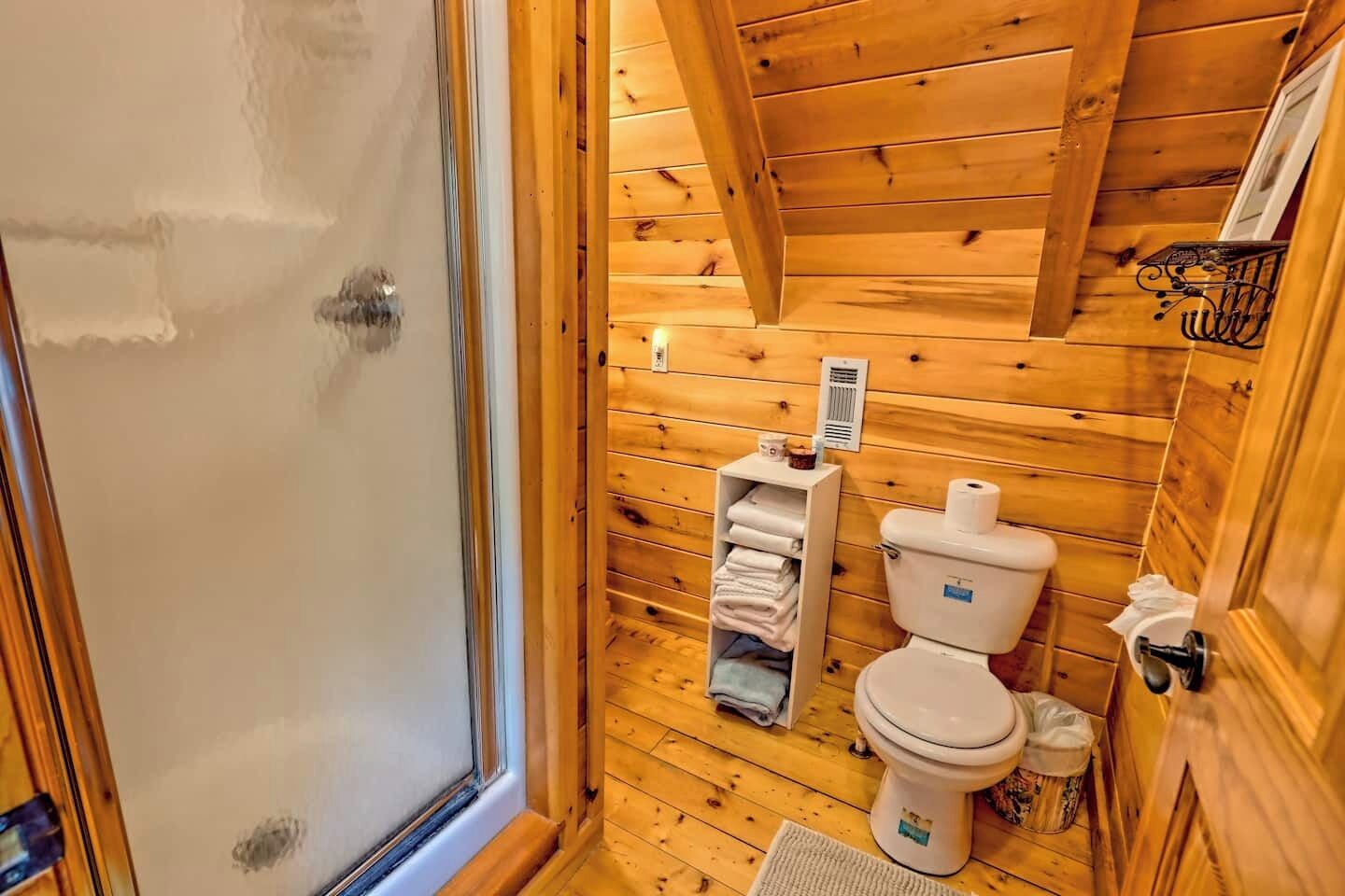 A small, wooden cabin bathroom with a toilet, a shelf with towels, and a window.