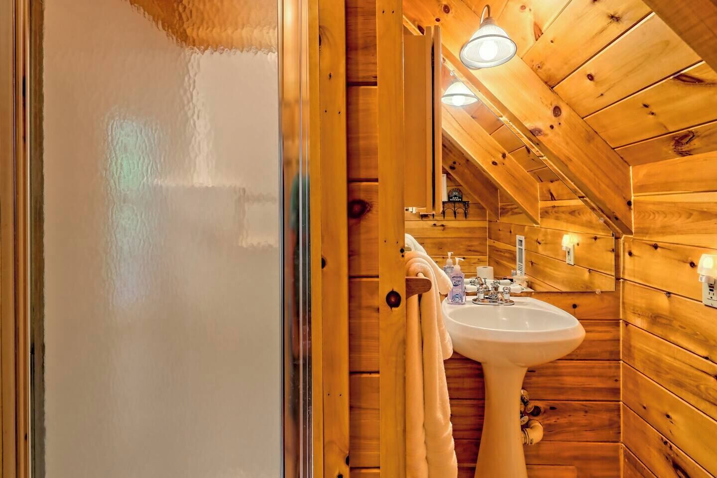 Baño de cabaña de madera con lavamanos blanco, una toalla colgada en la puerta y un espejo sobre el lavamanos.