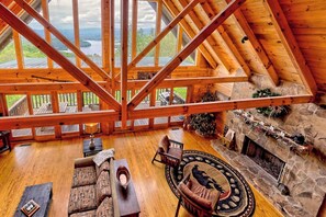 A spacious loft-style room with a high ceiling and large windows, a stone fireplace, and a view of a lake. - Brillant’s Bunk of the Blue Ridge Mountains by Chatuge Home Concierge | Lake & Mountain Views (Hayesville)
