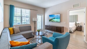 Una sala moderna con un sofá, sillones, una mesa de centro y un televisor. Se ve una cama con una mesita de noche y una lámpara al fondo.