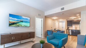 Una sala de estar moderna con un televisor de pantalla plana, un comedor con sillas azules y una cocina con armarios oscuros.