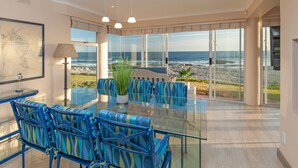 A modern dining area with a glass table and blue chairs, overlooking a rocky coastline.
