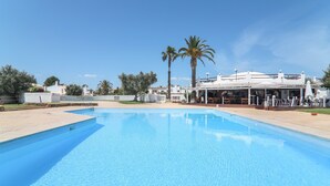 Una piscina con superficie de agua azul cristalina, rodeada por un edificio blanco con mobiliario exterior y palmeras.