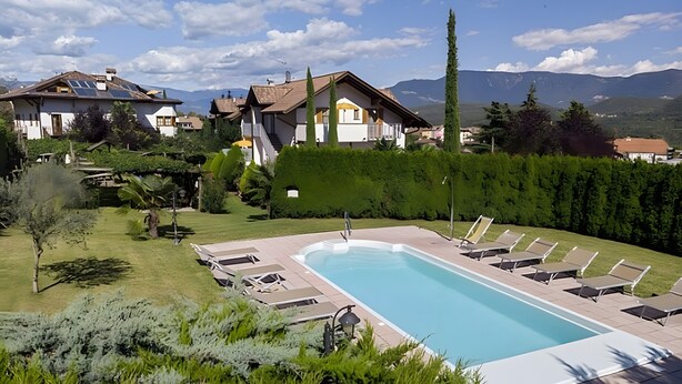 Una piscina con tumbonas, un jardín bien cuidado y casas al fondo.