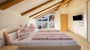 Un dormitorio con techo de madera, cama con ropa de cama estampada, mesita de noche, televisor montado en la pared y vista al exterior a través de una ventana.