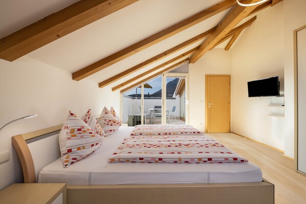 Une chambre avec un plafond en bois, un lit recouvert d’une literie à motifs, une table de chevet, un téléviseur fixé au mur et une vue sur l’extérieur par une fenêtre.