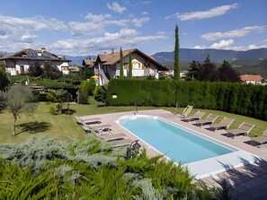 A swimming pool with lounge chairs, surrounded by well-maintained greenery and a view of houses and mountains.