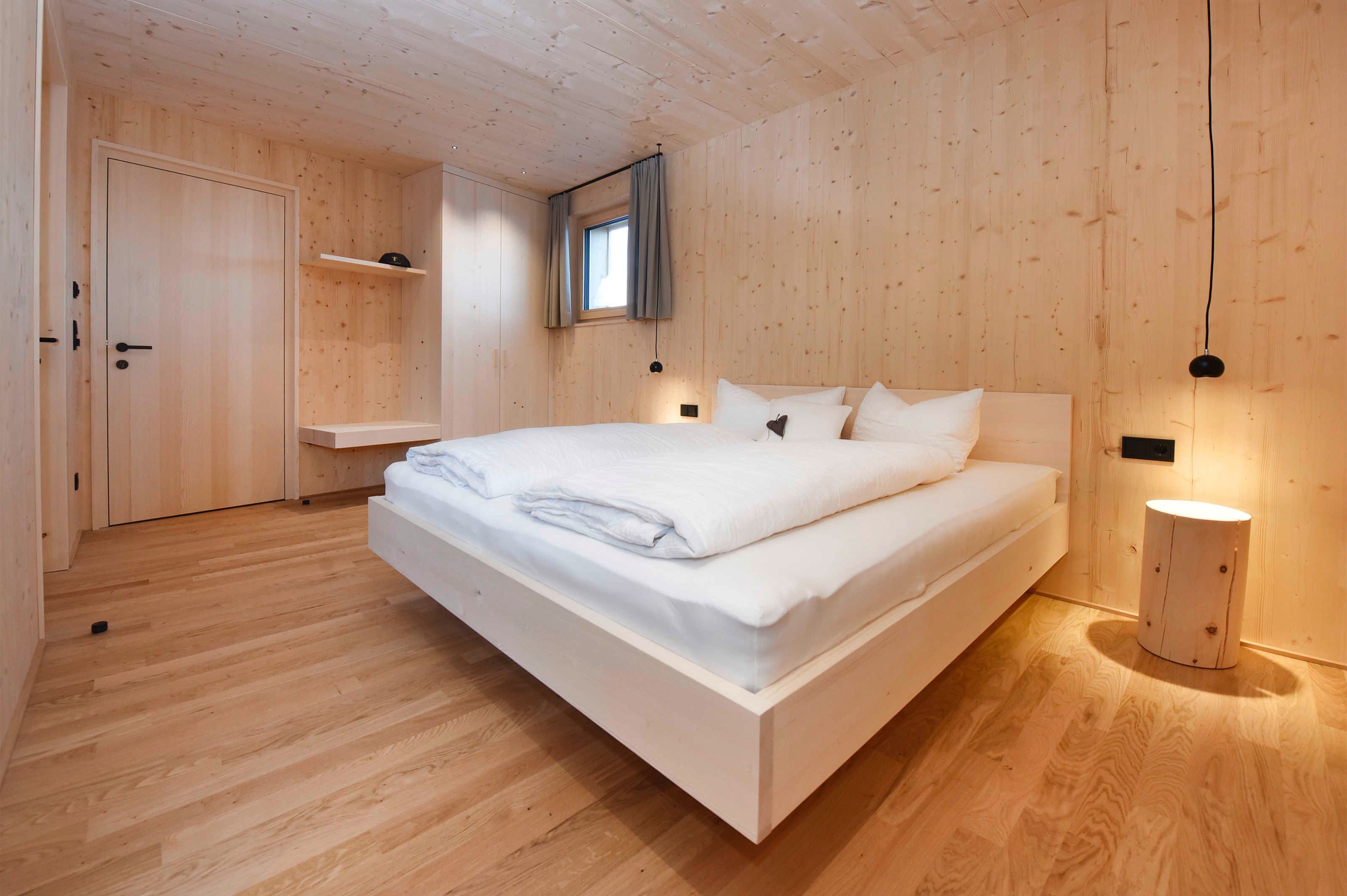 A wooden-paneled bedroom with a bed, a small window with curtains, and a bedside lamp.