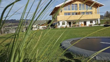 Une maison moderne en bois, dotée de grandes fenêtres, d’une cour verdoyante et d’un trampoline.