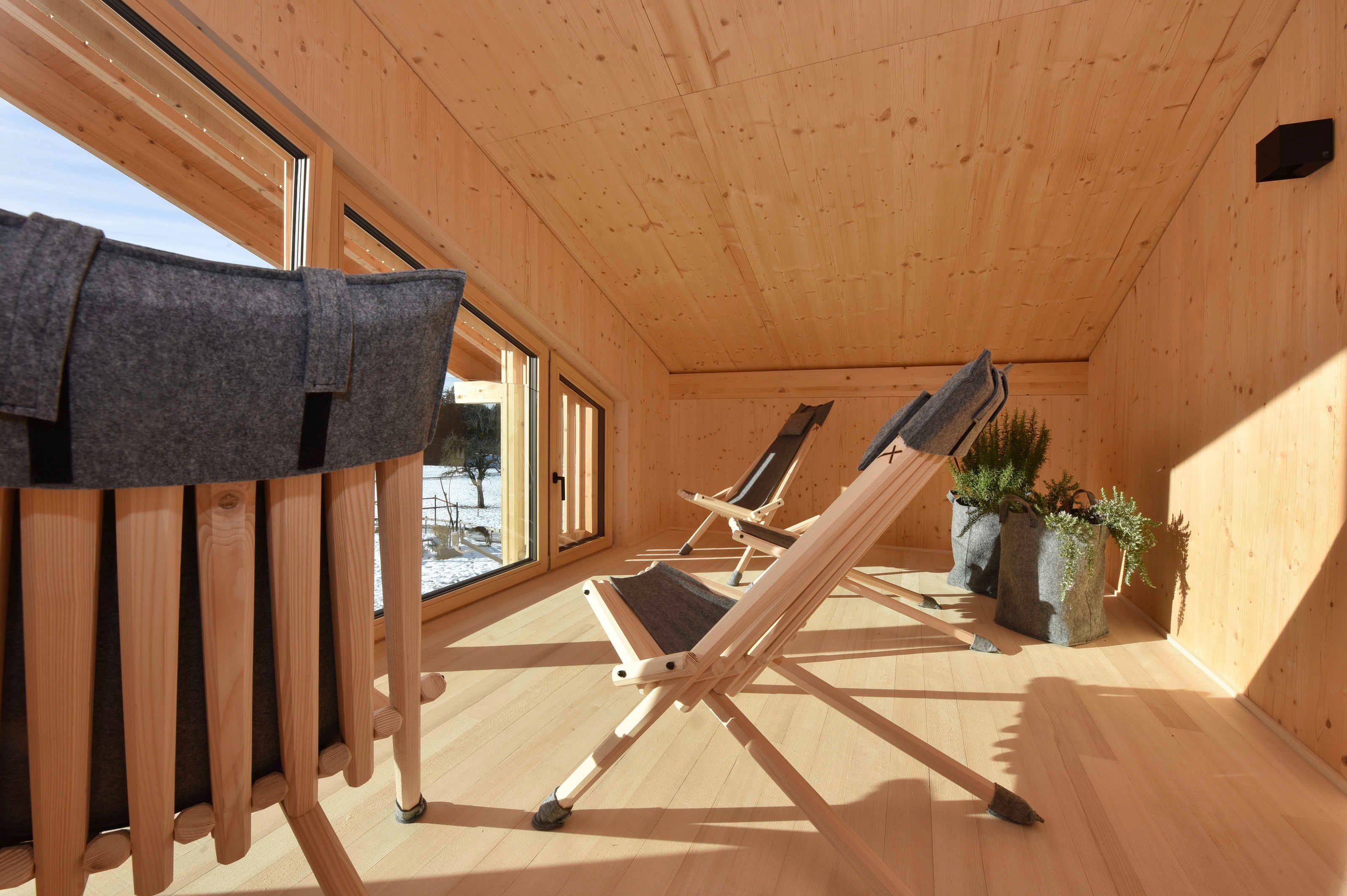 Un interior de madera con techo inclinado, dos sillones reclinables, una ventana con vistas de nieve y una planta en maceta.