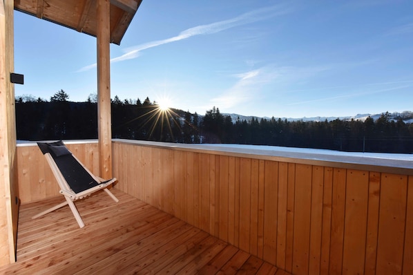 A wooden deck with a single lounge chair, a wooden railing, and a view of a snowy mountain range.