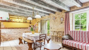 A rustic dining area with a brick wall, wooden ceiling beams, a round table with chairs, and a sofa with a patterned cover. - Holiday home  with garden (Villefranche-Du-Périgord)
