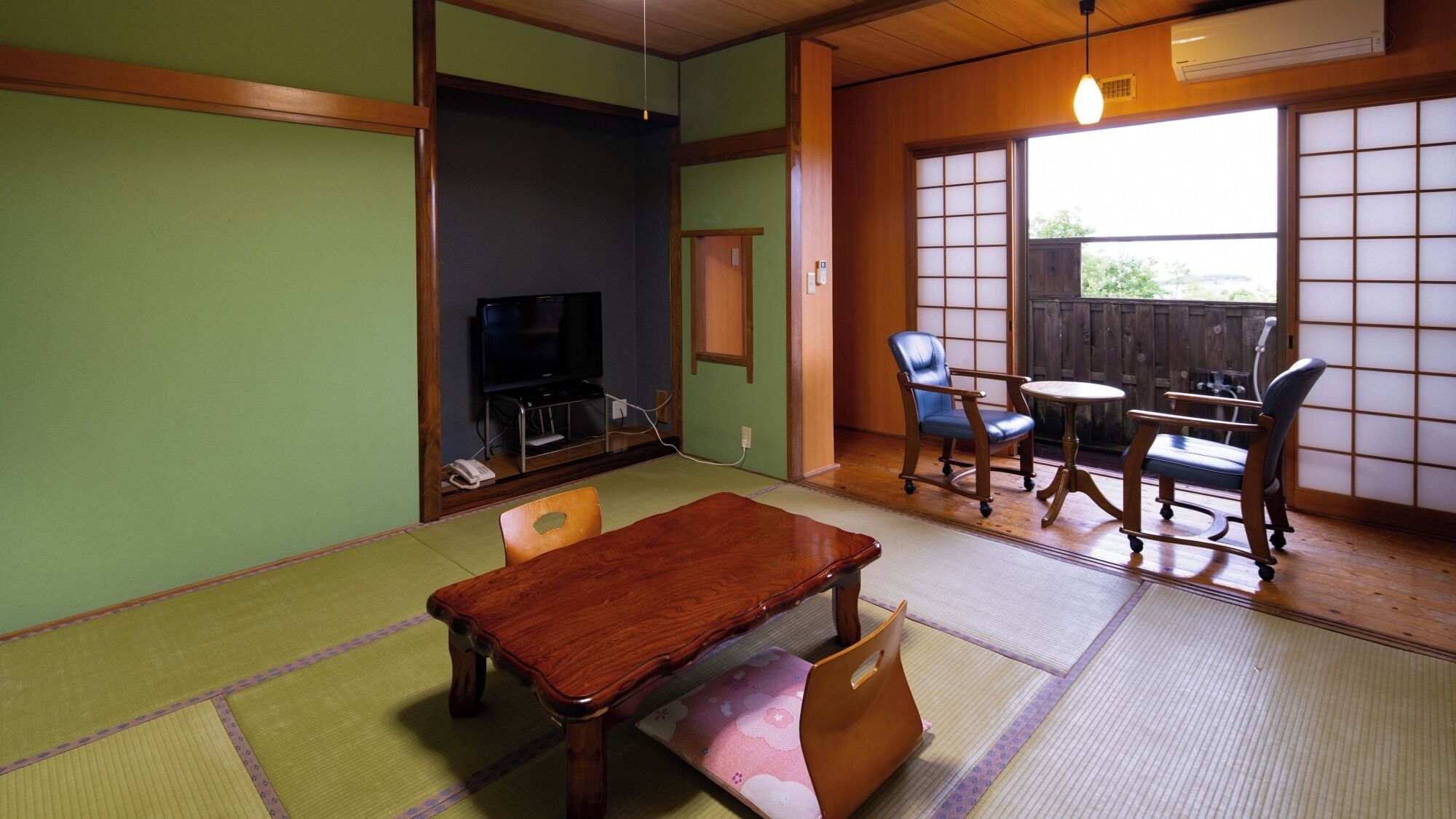 Ein traditioneller japanischer Raum mit Tatami-Boden, einem Holztisch, Stühlen, einem Fernseher und einer Schiebetür zu einem Außenbereich.