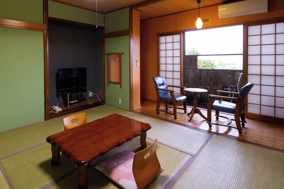 A traditional Japanese-style room with tatami flooring, a wooden table, chairs, a television, and a sliding door leading to an outdoor area.
